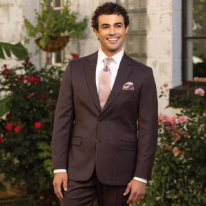 Man in a dark brown suit with a light pink tie and pocket square stands outdoors with flowers in the background, smiling warmly.