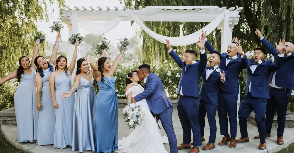 Bride and groom kiss beneath a white arch as bridesmaids in pale blue dresses cheer on and groomsmen in blue suits celebrate outdoors.