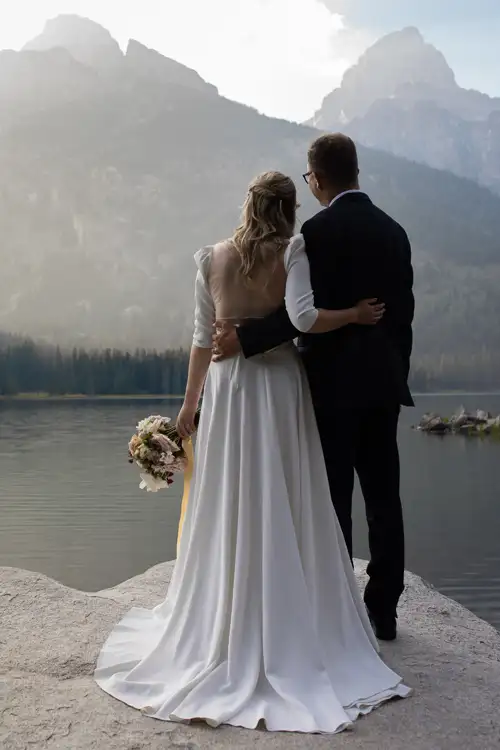 Bride in a white wedding gown and groom in a dark suit stand with arms around each other at the lake's edge, mountains rising in the background; the bride holds a bouquet.